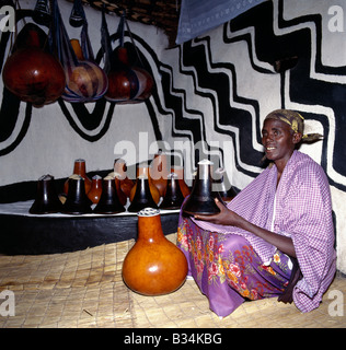 Uganda, Süd-Uganda, Mbarara. Bahima Frau mit ihrer Familie aus Holz Milchtöpfe (bekannt als Ekyanzi) und Kürbisse, die einen besonderen angehobenen Schritt in einer Ecke des Wohnzimmers gehalten werden. Häuser sind oft mit original afrikanischen Designs verziert. Die Bahima sind Banyarwanda sprechende Menschen leben in der Nähe der Grenze von Ruanda in Süd-Uganda. Sie halten lange gehörnten Ankole Rinder, eine afrikanische Taurin Rasse mit Ursprung vor der Einschleppung der buckligen Rücken oder Zebu Rinder am Horn von Afrika während der menschlichen Invasionen aus Arabien im siebten Jahrhundert v. Chr. zurückgeht. Stockfoto