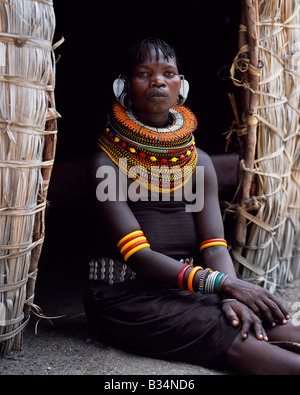 Kenia, Northern Frontier District Loyangalani. Eine Turkana-Frau, in der Regel tragen viele Schichten von Perlen Halsketten und eine Reihe von geringelten Ohrringe mit einem Paar blattförmigen Earrrings an der Vorderseite sitzt am Eingang zu ihrer Hütte. Stockfoto