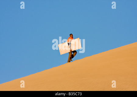 Eine Mann klettert Sanddüne mit seinem Brett Sandboarding auf Sanddünen in der Nähe von Swakopmund in Namibia Afrika gehen Stockfoto