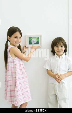 Mädchen stehen neben jüngeren Bruder, hängen gerahmtes Bild, beide lächelnd in die Kamera Stockfoto