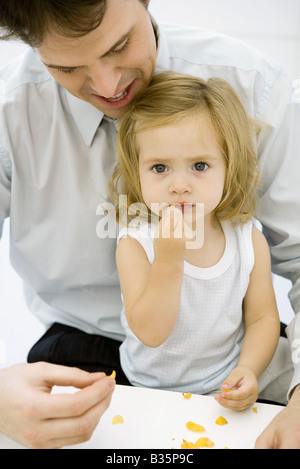 Junge Mädchen essen ein Stück von Getreide, sitzen in des Vaters Schoß, Mann, ihr zu helfen Stockfoto