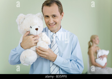 Vater holding Teddybär, lächelnd in die Kamera, Frau halten Baby im Hintergrund Stockfoto