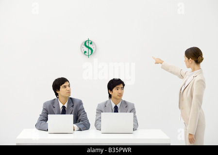 Zwei männliche Büroangestellte sitzen am Schreibtisch, weibliche Betreuer zeigen Uhr mit Dollarzeichen Stockfoto