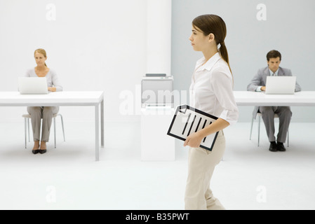 Weibliche Büroangestellte mit Dokument unter Arm, im Hintergrund arbeitenden Kolleginnen und Kollegen Stockfoto