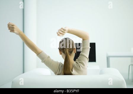Junge Frau sitzt im Sessel, Arme ausgestreckt, Rückansicht Stockfoto