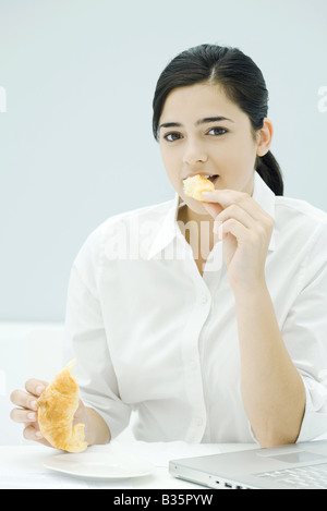 Junge Frau Essen Croissant, sitzen am Schreibtisch, Blick in die Kamera Stockfoto