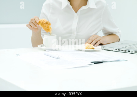 Frau eintauchen Croissant in Kaffee, unaufgeräumten Schreibtisch, beschnitten Ansicht Stockfoto