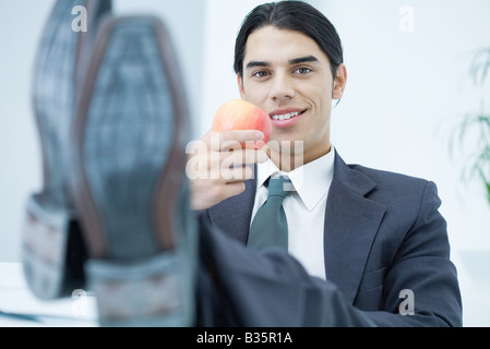 Professionelle Jüngling mit Füßen sitzend, hält Apfel und lächelt in die Kamera Stockfoto