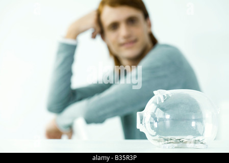 Transparente Sparschwein sitzend am Tisch, jungen Mann im Hintergrund, die Kopf haltend Stockfoto