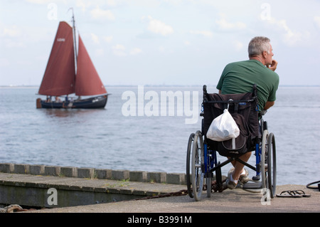 Männliche Person im Rollstuhl genießt die Aussicht auf die Oosterschelde, wenn ein Segelschiff vorbei. Zeeland. Niederlande. Stockfoto