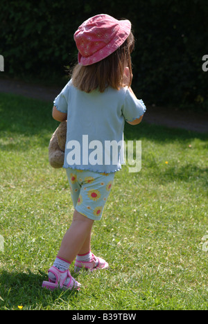 Rückansicht eines kleinen Mädchens halten einen Teddybär und Shorts und ein Sonnenhut zu Fuß entfernt Stockfoto