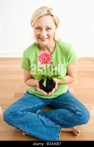 Porträt von lächelnden Erwachsenen blonde Frau sitzt auf Etage mit rosa Gerber Daisy Pflanze Stockfoto