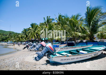 Fischerboote Playa Principal Zihuatanejo Mexiko | ZIHUATANEJO, Mexiko – Panga Fischerboote sitzen am Playa Principal am Strand, ihre Außenbordmotoren sind nach der Landung am Morgen aus dem Wasser gekippt. Diese kleinen offenen Boote bilden das Rückgrat der lokalen ribereño-Fischerei, die hauptsächlich Huachinango und andere Schnapper-Arten anlegt, die direkt am Strand verkauft werden. Kokospalmen und niedrige Hügel umrahmen die Bucht dahinter. Stockfoto