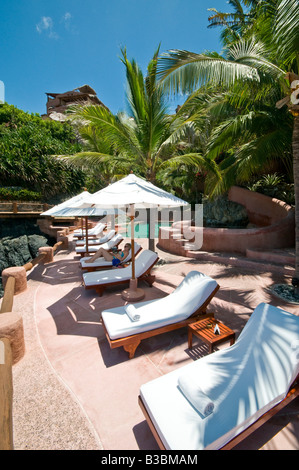 La Casa Que Canta Resort Zihuatanejo Mexiko | ZIHUATANEJO, Mexiko – Ein Gast entspannt sich zwischen den weißen Sonnenschirmen am Pool im La Casa Que Canta, einem Terrassenresort oberhalb der Bucht von Zihuatanejo in Guerrero, Mexiko. Die strohgedeckten Palapaüberdachungen und die terrakottafarbene Architektur hinter dem Pool sind charakteristisch für den pazifischen Küstenresort-Stil der Region, der sich in den palmenbedeckten Hügel einfügt. Stockfoto