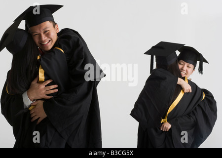 Junge männliche Absolvent umarmt weibliche Absolventin und zwei weibliche Absolventen umarmt im Hintergrund Stockfoto