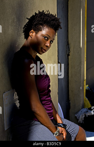 Schöne schwarze braune afroamerikanische Frau im lila Hemd sitzen gegen eine Wand im freien Stockfoto