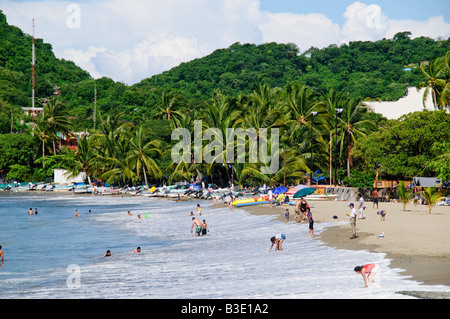 Playa Principal Beach Zihuatanejo Mexiko / ZIHUATANEJO, Guerrero, Mexiko – der Playa Principal Beach ist eine lebendige Szene, in der Menschen das Wasser genießen und zahlreiche kleine Boote auf den Sand gezogen werden. Üppige Palmen und grüne Hügel umgeben diesen beliebten Küstenort. Stockfoto