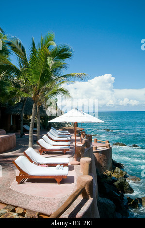 La Casa Que Canta Resort Zihuatanejo Mexiko | ZIHUATANEJO, Mexiko - Liegestühle und ein Sonnenschirm blicken auf das offene Wasser im La Casa Que Canta, einem Resort an der Klippe mit Blick auf den Pazifik südlich der Zihuatanejo Bay. Die von Kokospalmen und einem strohgedeckten Palapadach eingerahmte Terrasse ist typisch für das gestufte, auf den Ozean ausgerichtete Design, das Boutique-Hotels an der Pazifikküste Mexikos von größeren Strandresorts unterscheidet. Stockfoto
