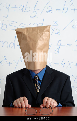 Mann in einem Anzug mit einer braunen Papiertüte auf dem Kopf sitzt hinter seinem Schreibtisch Stockfoto