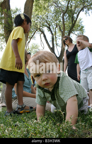 acht Monate altes Baby junge kriechen auf dem Rasen im Park mit Familie und Freunden stehen hinter ihm Stockfoto