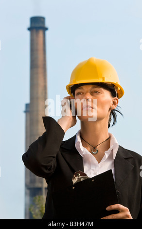 Eine schöne dunkelhaarige Frau trägt einen Schutzhelm und sprechen über ihr Mobiltelefon im Stand vor einer Fabrikgebäudes Stockfoto