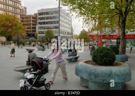 Asiatische Mutter ihr Baby in einem Kinderwagen schieben, über Marktschreier Pool Platz, Sheffield. Eine andere Mutter ist auf Sitzbank Baby füttern. Stockfoto