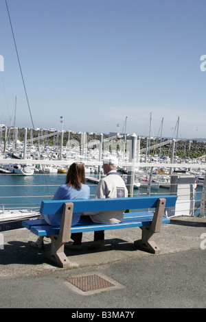 Nautische Szene mit älterer Mann und jüngere Frau sitzen zusammen auf Sitzbank mit Blick auf Hafen mit Booten & Yachten vor Anker Stockfoto