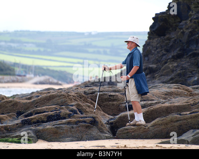 50er Jahre plus Mann auf Felsen mit Blick auf das Meer mit Wanderstöcken für Unterstützung. Stockfoto