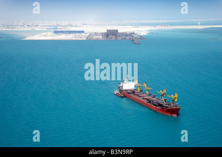 Eine Frachtschiff liegt vor Anker von der Küste von Bahrain in Reich Blau des Meeres mit Schwerindustrie im Hintergrund. Luftaufnahme. Stockfoto