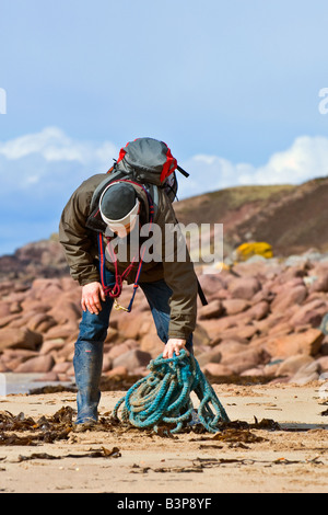 Mann mit Rucksack auf dem Rücken aufrollen etwas blauem blauen Seil, das am Slaggan Strand, Wester Ross, Schottland an den Strand gespült worden ist Stockfoto