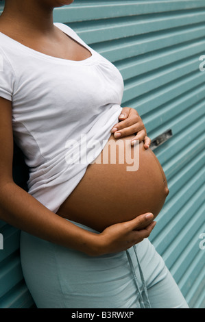 Schwanger unverheiratete schwarzen Teenager alleinerziehende Mutter Mädchen allein, UK, halten Sie ihren Bauch, stützte sich auf blauen Metall Garagentor Stockfoto