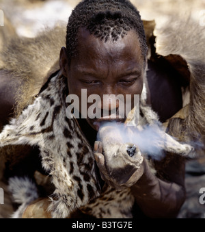 Tansania, Norden von Tansania, Lake Eyasi. Hadza Jäger tragen ein Genet Katze Haut Cape raucht Cannabis aus einem rohen Stein Rohr in Leder ummantelt. Die Hadzabe sind eine 1000-Seelen-Gemeinde von Jägern und Sammlern, die in der Lake Eyasi-Becken seit Jahrhunderten gelebt haben. Sie sind einer von nur vier oder fünf Gesellschaften in der Welt, die immer noch in erster Linie von der Wildtiere Leben verdienen. . Stockfoto