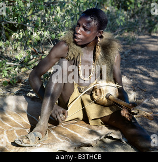 Tansania, Arusha, Lake Eyasi. Sitzen auf der Haut eine große Kudu, singt Hadza Jugendlicher ein Pavian Haut Cape tragen, begleitet von seinem zweisaitiges Musikinstrument. Die Hadzabe sind eine 1000-Seelen-Gemeinde von Jägern und Sammlern, die in der Lake Eyasi-Becken seit Jahrhunderten gelebt haben. Sie sind einer von nur vier oder fünf Gesellschaften in der Welt, die immer noch in erster Linie von der Wildtiere Leben verdienen. Stockfoto