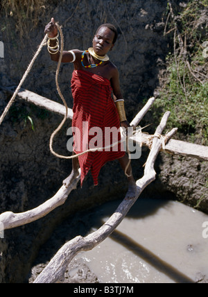 Tansania, Norden von Tansania, Manyara. Eine junge Frau Datog zieht das Wasser aus einem tiefen Brunnen, prekär auf Baumstämmen balancieren. Sowohl Männer als auch Frauen arbeiten Brunnen um ihr Vieh zu Wasser. Sie trägt einen traditionellen gewickelten Messing-Kette und Armulet, und ihr Gesicht hat umfangreiche erhöhten Scarification Runde Augen, eine Verschönerung, die Frauen eigen ihres Stammes. Die Datog (ihren Maasai Nachbarn als die Mang'ati und der Iraqw als Babaraig bekannt) Leben im Norden von Tansania und sind vor allem Hirten. Stockfoto