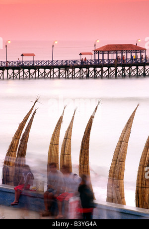 Peru, La Libertad, Huanchaco. Totora (Schilf) Boote sind entlang des Strandes, mit dem hölzernen Pier im Hintergrund bei der Fischerei Huanchaco, in der Nähe von Trujillo im Norden Perus gestapelt. Stockfoto