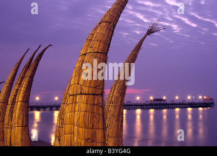 Peru, La Libertad, Huanchaco. Totora (Schilf) Boote sind entlang des Strandes, mit dem hölzernen Pier im Hintergrund, die Fischerei Dorf von Huanchaco im Norden Perus gestapelt. Die Boote genannt Caballitos de Totora (kleine Pferde Schilf) sind das traditionelle Handwerk der lokalen Fischer. Stockfoto