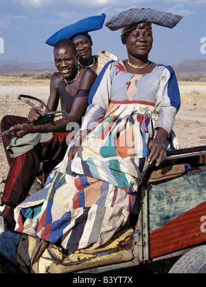 Namibia, Damaraland, Palmwag. Ein Herero-Mann und zwei Frauen fahren Sie nach Hause in einem Eselskarren. Die aufwändige Kleider und einzigartigen Hut Stile der Herero-Frauen können auf deutsche Missionare des 19. Jahrhunderts zurückführen, die Ausnahme, was sie eine unbescheidene Form von Kleid unter dem Stamm als nahm. Sie bestand darauf, dass die Herero Frauen ihre eigenen unpraktisch Stil der viktorianischen Kleider anzunehmen. Im Laufe der Jahre entstanden bunte Kleidern der Herero als enorme Krinolinen über fünf Unterröcken getragen. Stockfoto