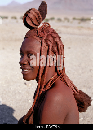 Namibia, Kaokoland, Purros. Ein Himba-Frau in traditioneller Kleidung. Ihr Körper glänzt aus einer Mischung von rotem Ocker, Butterfett und Kräutern. Ihr langes Haar ist in der traditionellen Himba Weise gestaltet und mit einem Kopfschmuck aus Lammfell, genannt Erembe gekrönt wird. Die Himba sind Bantu Herero sprechende Nomaden, die in der rauen, trockenen aber krass schöne Landschaft entlegenen Nordwesten Namibias Leben. Stockfoto