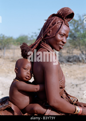 Namibia, Kaokoland, Opuwo. Himba Mutter und Kind reiten nach Hause auf einem Esel. Ihre Körper glänzen aus einer Mischung von rotem Ocker, Butterfett und Kräutern. Lange Haare der Frau ist in der traditionellen Himba Weise gestaltet und wird gekrönt mit einem Kopfschmuck aus Lammfell, genannt Erembe. Die Himba sind Bantu Herero sprechende Nomaden, die in der rauen, trockenen aber krass schöne Landschaft entlegenen Nordwesten Namibias Leben. Stockfoto