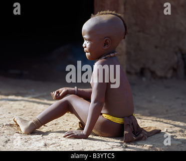 Namibia, Kaokoland, Epupa. Ein kleines Mädchen sitzt außerhalb ihrer Familie zu Hause. Ihr Körper ist mit einer Mischung aus rotem Ocker, Butterfett und Kräutern bestrichen. Himba Mütter schneiden ihre Kinder Haare in einigen auffälligen Arten. Das Kind trägt eine Runde Halskette, genannt Ombwari und ein Perlen Gürtel aus welche hängen ihre bescheidenen vorne und hinten Leder Bekleidung. Die Himba sind Bantu Herero sprechende Nomaden, die in der rauen, trockenen aber krass schöne Landschaft entlegenen Nordwesten Namibias Leben. Stockfoto