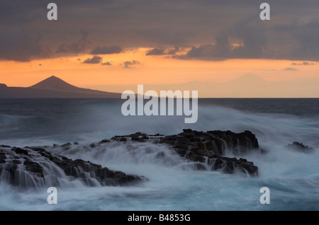 Wellen brechen über Felsen bei Sonnenuntergang am El Confital, Gran Canaria. Galdar (links) und den Teide auf Teneriffa in der Ferne. Stockfoto