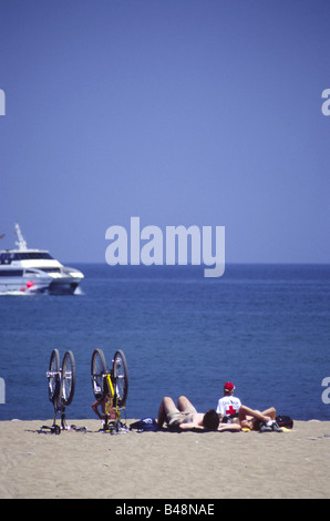 Sonnenbaden am Strand von Barceloneta liegen neben Männer umgedrehten Fahrräder, träge beobachten eines vorbeifahrenden Bootes, Barcelona, Spanien Stockfoto