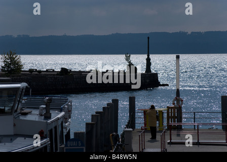 Sonnenlicht glitzert ab Neuenburger See. Der Port de Neuchâtel steht im Vordergrund; einer der Fähren ist angedockt. Frau am Pier. Stockfoto