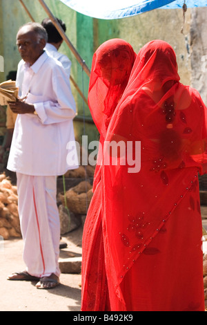 Frauen in roten Saris und Schleier besuchen einen Straßenmarkt in Mysore, Indien. Im Hintergrund steht ein Mann in einem weißen Anzug. Stockfoto