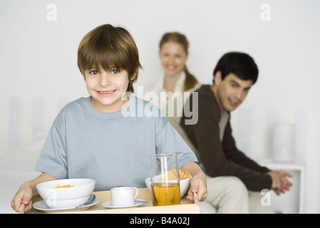 Kleiner Junge mit Frühstück auf Tablett, Eltern sitzen am Bett im Hintergrund Stockfoto