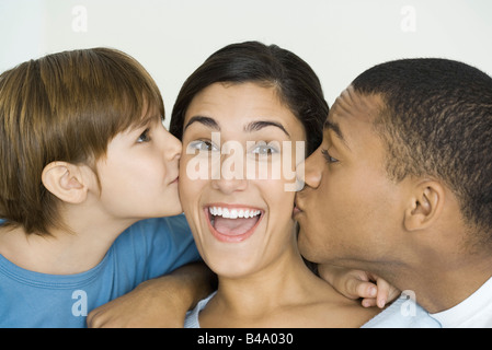 Frau in die Kamera Lächeln, als Ehemann und Sohn ihre Wangen küssen Stockfoto