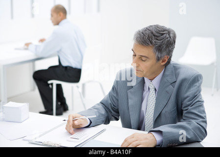 Professionelle Mann sitzt am Schreibtisch, mit Taschenrechner, Kollege im Hintergrund Stockfoto