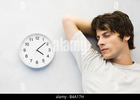 Junger Mann lehnte sich gegen die Wand neben Uhr, Augen geschlossen Stockfoto