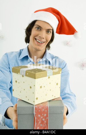 Junger Mann trägt Weihnachtsmütze, Weihnachtsgeschenke, durchhalten und Lächeln in die Kamera Stockfoto