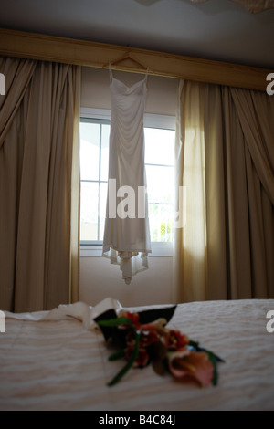 Hochzeitskleid von einem Fenster mit Blumen auf dem Bett im Schlafzimmer hängen Stockfoto